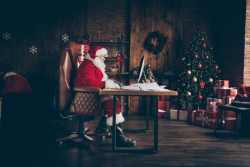 Profile side full length photo focused grey hair santa claus sit table work computer check mail wish list letters wear red costume boots in house indoors with christmas x-mas ornament - Powered by Adobe