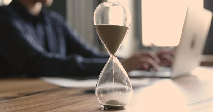 Businesswoman work typing on laptop hands and pc view sit at workplace desk, on foreground close up hourglass showing passage of time. Billable hours, completion of work on time, time is money concept