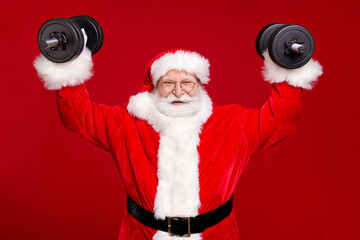 Photo of retired grandpa grey beard hold easily lift dumbbells show strength raise arms smile wear costume leather belt gloves coat spectacles headwear isolated red color background