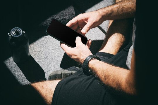 Close up of a man holding his mobile phone with an empty screen, while taking a break in between exercises inside a gym.
