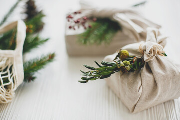 Zero waste christmas holidays. Stylish christmas gift wrapped in linen fabric with green branch and reusable shopping bag with green spruce on rustic wooden background. Sustainable lifestyle