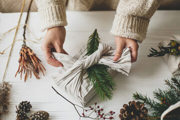 Hands decorating stylish christmas gift in linen fabric with green fir branch on white rustic table...