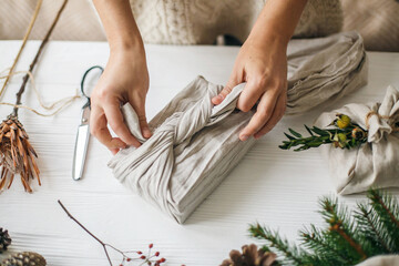 Hands wrapping christmas gift in linen fabric in furoshiki style on white rustic table with natural fir, herb, pine cones and scissors. Female preparing plastic free christmas present
