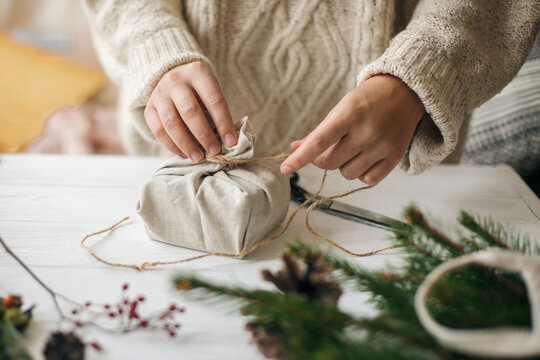 Hands wrapping stylish christmas gift in linen fabric on white rustic table with fir, pine cones, scissors, twine. Female in cozy sweater preparing plastic free christmas present, zero waste