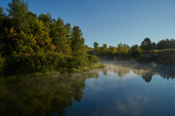 Fototapeta premium Autumn time. Dawn over the river in a misty, brooding haze. Beautiful view of the forest and river, covered with fog in the early morning. The sun's rays of light. September.