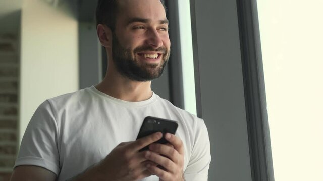Young happy man standing near window indoors at home while using mobile phone