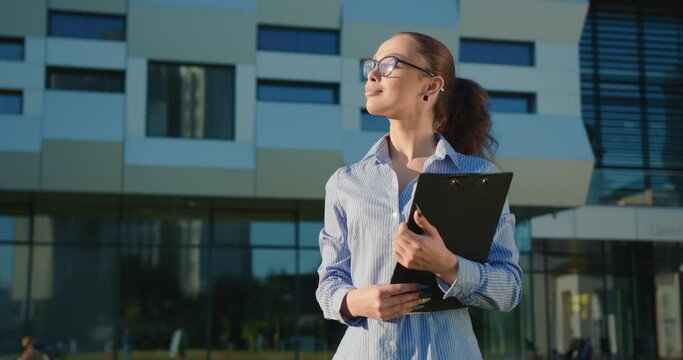 A businesswoman is walking near the business center. She is smiling and holding a folder in her hands. She's wearing a shirt. The sun is shining. 4K