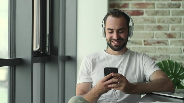 Young pleased man listening music with headphones indoors at home while holding mobile phone