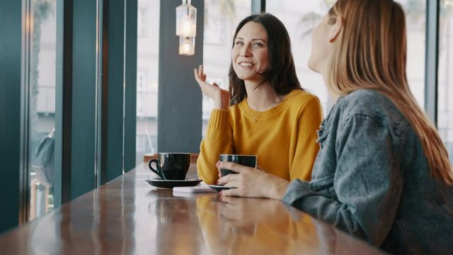 Two young women sitting at cafe table chatting and laughing. Female friends meeting at coffee shop on a weekend. 
