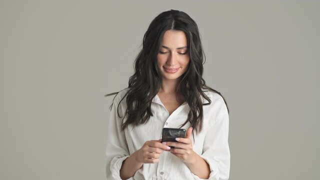A dreaming young woman is typing on her smartphone standing isolated over gray background