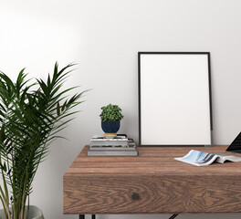 Vertical black frame above a walnut wood desk with books with a small blue vase with plants to one side, a magazine and a palm houseplant in a concrete vase to the side in front of a white wall