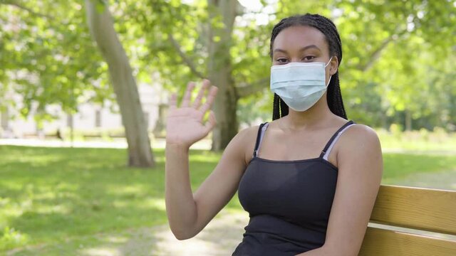A young black woman in a face mask waves at the camera and sits on a bench in a park on a sunny day