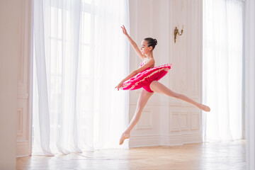 A little ballerina in a bright pink tutu is dancing in a beautiful white hall, froze in a jump