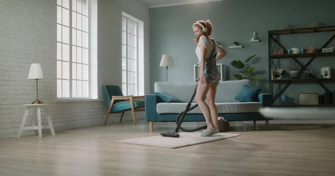 Young positive caucasian woman funnily dancing while cleaning the house. Cute girl enjoys tidying her home, using vacuum cleaner and listening to music - happy people 4k footage