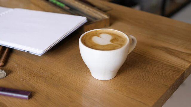 male waiter puts cup of coffee with milk heart picture on wooden table with different art accessories in cafe closeup slow motion