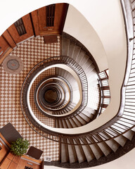 Brown round spiral staircase with white walls black railings with patterns and checkered floor down view