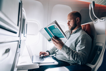 Fototapeta premium Serious man typing on laptop and reading book in aircraft