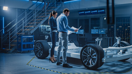 Fototapeta na wymiar Auto Industry Design Facility: Male Chief Engineer Shows Car Blueprints Female Software Design and Integration Engineer. Electric Vehicle Platform Chassis Concept Has Wheels, Engine and Battery 