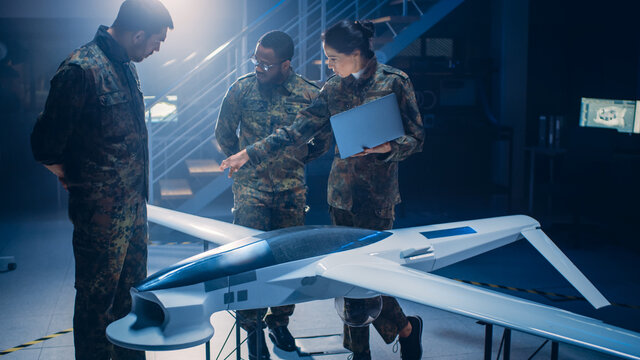 Army Aerospace Engineers Work On Unmanned Aerial Vehicle / Drone. Uniformed Aviation Experts Talk, Using Laptop. Industrial Facility with Aircraft for: Surveillance, Warfare Tactics, Air Strike