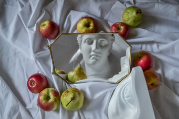 Ancient greek sculpture white plaster marble head of diana and eye of david reflected in mirror in frame against background of white drapery fabric with folds with red apples and green pears