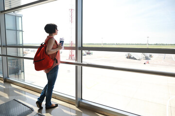 Obraz na płótnie Canvas Brunette caucasian woman indoors airport in gate and looking through the window on the aircrafts preparing for departure.