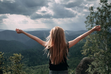 Young blond girl with hands up enjoing view of the evergreen taiga forest hills on rainy day. Stolby Nature Reserve in Krasnoyarsk, Russia.