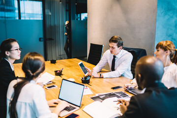 Group of diverse male and female colleagues sitting at conference table with mockup laptop discussing web project during collaborative meeting, multiracial persons teamworking on ideas in company