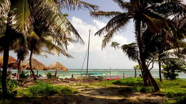 Costa Maya / Mexico - December 2019: People relaxing and playing volleyball on a sunny beach in the Mahahual village. Palm thatch sun umbrellas, sun chairs, catamaran and palm trees in the wind
