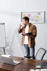 young businessman in casual clothes drinking coffee and holding notebook while standing in office