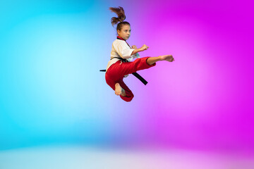 Fototapeta na wymiar In jump. Karate, taekwondo girl with black belt isolated on gradient background in neon light. Little caucasian model, sport kid training in motion and action. Sport, movement, childhood concept.