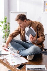 Selective focus of shocked businessman holding notebook and papers near digital devices on office table