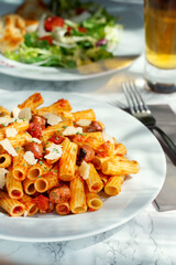 Plate of rigattone pasta with chorizo ​​and cheese on white plate and gray napkin. In the background beer and salad