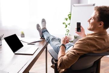 Selective focus of young businessman holding coffee cup and cellphone near laptop on office table