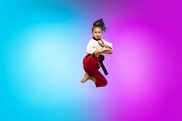 Fototapeta na wymiar In jump. Karate, taekwondo girl with black belt isolated on gradient background in neon light. Little caucasian model, sport kid training in motion and action. Sport, movement, childhood concept.