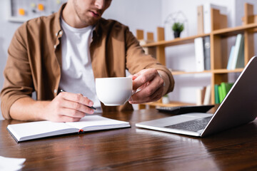 Selective focus of businessman holding cup of coffee while working with notebook near laptop