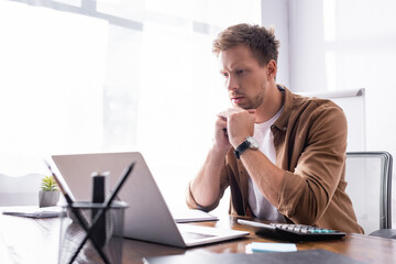 Naklejka premium Selective focus of young businessman looking at laptop near calculator and stationery in office