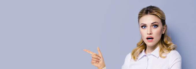 Portrait of excited surprised blond businesswoman with opened mouth, pointing something or copy space area for some imaginary, slogan or text. Success in business concept. Grey color background.