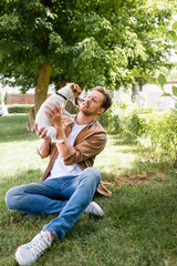 Naklejka na ściany i meble young man in casual clothes holding jack russell terrier dog while sitting on green grass