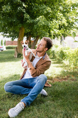 man in brown shirt and jeans holding jack russell terrier dog while sitting on lawn in park