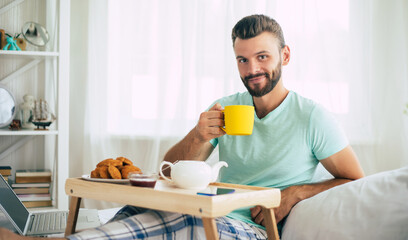 Happy young bearded man is sitting on his bed and has his breakfast with tea or coffee