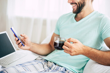 Close up photo of young man with a laptop and smartphone drinks coffee in bedroom.