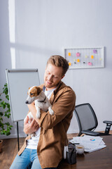 businessman in casual clothes sitting on office desk and holding jack russell terrier dog