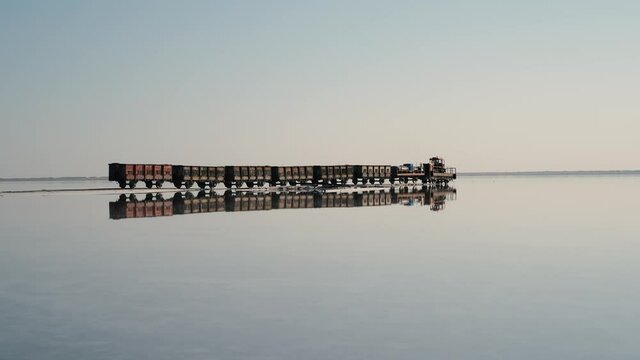 An old train travels on a railway laid in the water across a salt lake. Extraction of salt in Lake Bursol. Russia. September 2020