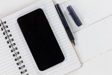View of a black smartphone, a notebook and a black pen on plain white background