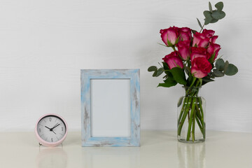 View of a picture frame and a clock with pink roses placed in a glass vase on plain white background