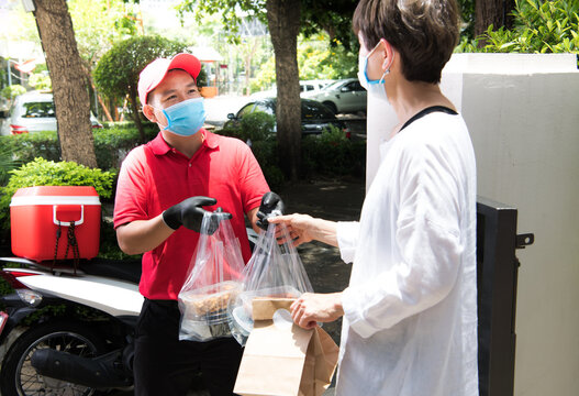 Asian Delivery Man Wearing Face Mask And Gloves In Red Uniform Delivering Bag Of Food And Drink To Recipient During COVID-19 Outbreak