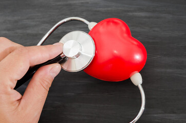 Doctor with a stethoscope examines a red heart on a dark wooden background. Close-up.