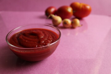 red tomato sauce in a glass saucepan plate next to the back are vegetables tomatoes and onions on a pink background copy space
