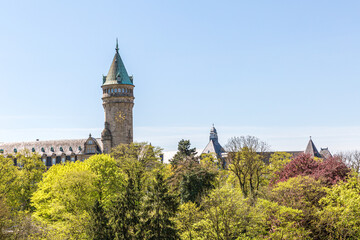 Fototapeta na wymiar Trees with green leaves and the banking museum with its tower and clock in the background, sunny spring day with a blue sky in Luxembourg city