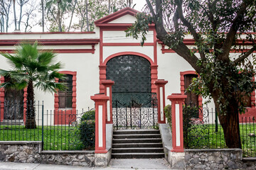 Facade of a white building with red details, with black wrought iron fence, a small concrete staircase and a metal gate in Colomos Park, cloudy day in Guadalajara Jalisco, Mexico